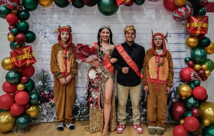 Four young people pose in front of a festive holiday backdrop with red, green, and gold balloons. The person in the center wears a crown, sash, and formal dress while holding a bouquet, flanked by two people in reindeer costumes and one in a black shirt with a sash. Christmas-themed decorations surround them.