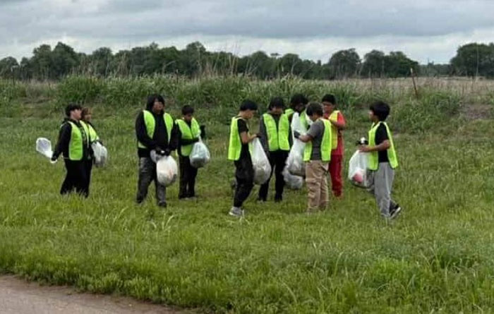 A group of students wearing reflective safety vests collect trash in a grassy field during a community roadside cleanup on a cloudy day.