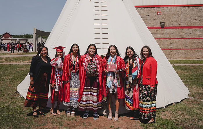 Six women pose in front of a large white teepee, with four graduates in red gowns and traditional patterned clothing smiling outdoors on a sunny day.