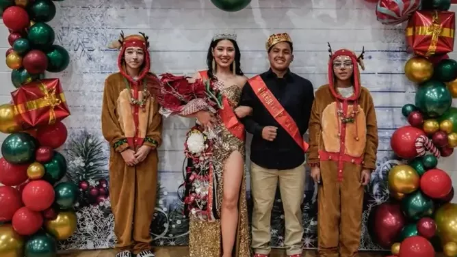 Four young people pose in front of a festive holiday backdrop with red, green, and gold balloons. The person in the center wears a crown, sash, and formal dress while holding a bouquet, flanked by two people in reindeer costumes and one in a black shirt with a sash. Christmas-themed decorations surround them.