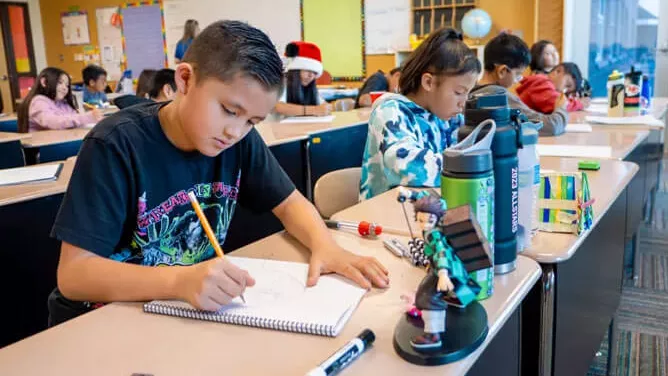 Elementary school students sit at desks in a classroom, focused on writing and drawing in notebooks, with water bottles and school supplies on their desks.