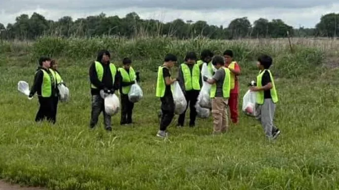 A group of students wearing reflective safety vests collect trash in a grassy field during a community roadside cleanup on a cloudy day.