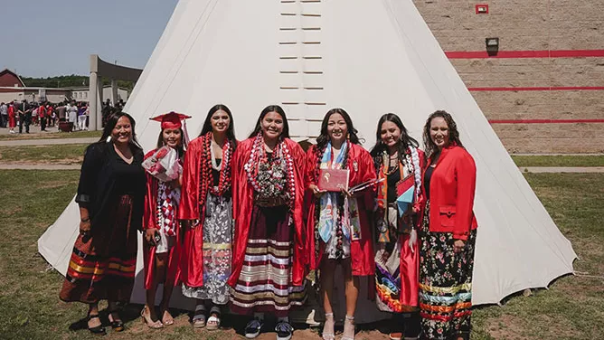 Six women pose in front of a large white teepee, with four graduates in red gowns and traditional patterned clothing smiling outdoors on a sunny day.
